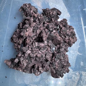 May include: A cluster of dark, textured rocks with a reddish-brown hue. The rocks have a rough, uneven surface and are arranged in a compact formation. The image is taken from above, showing the rocks against a light blue background.