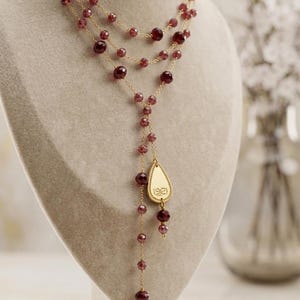 Puede incluir: Un collar de varias capas con cadenas doradas y cuentas de color burdeos. El collar presenta un colgante en forma de lágrima con un diseño grabado. El collar se exhibe en un maniquí.