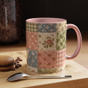 May include: A ceramic mug with a patchwork quilt design. The mug features a pink interior and handle, and the exterior is decorated with squares of floral and gingham patterns in pastel colors. A spoon and a macaron are on a wooden surface.