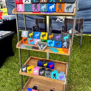May include: A display of Bigfoot-themed accessories, including seat covers and coasters, in various colors and patterns. The items are arranged on a multi-tiered wooden shelf, with some featuring the text "Buck It" and "Stay Wild".