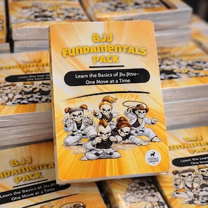 May include: A deck of cards titled "BJJ Fundamentals Pack" with the text "Learn the Basics of Jiu-Jitsu - One Move at a Time." The cover features cartoon skeletons in martial arts uniforms. The background is yellow with radiating lines.