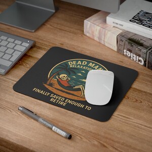 Peut inclure: Tapis de souris rectangulaire noir avec le texte "DEAD MAN RELAXATION" et "FINALLY SAVED ENOUGH TO RETIRE". Le graphisme représente un squelette se relaxant dans un hamac sur une plage. Une souris d'ordinateur blanche est posée sur le tapis.