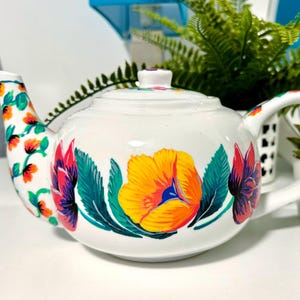 May include: White ceramic teapot with a floral design. The teapot features a large yellow flower with blue accents, surrounded by red and purple flowers and green leaves. The spout and handle are also decorated with flowers.