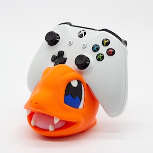 Puede incluir: Un soporte para mando naranja con temática de Charmander con un mando Xbox blanco encima. El soporte presenta el rostro del personaje Pokémon, con ojos azules y una lengua rosa. El mando tiene botones negros y botones de acción de colores.