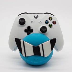 Puede incluir: Un mando blanco de Xbox descansa sobre una esfera azul adornada con gafas de sol a rayas blancas y negras. El mando tiene joysticks negros, una cruceta y botones de acción de colores.