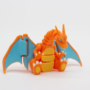 Puede incluir: Figura de Charizard impresa en 3D, naranja y azul. La figura presenta un diseño detallado con alas naranjas, vientre amarillo y alas azules. Este personaje es de la franquicia Pokémon.