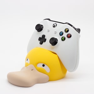 Puede incluir: Un mando blanco de Xbox descansa sobre un soporte para mando con forma de pato amarillo y beige. El mando tiene botones negros y botones de acción de colores. El soporte para pato tiene un pico grande de color beige y está diseñado para sujetar el mando.