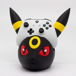 Puede incluir: Un accesorio de juego con temática Umbreon en negro y amarillo con un mando de Xbox blanco encima. La figura de Umbreon tiene ojos rojos y un anillo amarillo. El mando tiene botones negros y una cruceta.