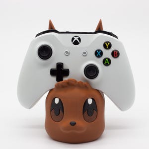 Puede incluir: Un mando blanco de Xbox descansa sobre un soporte marrón con forma de personaje de dibujos animados. El mando tiene botones negros y botones de acción de colores. El soporte tiene ojos grandes y orejas pequeñas. El fondo es blanco liso.