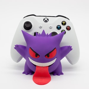 Peut inclure: Une figurine Gengar violette avec des yeux rouges et une langue rouge, devant une manette Xbox blanche. La manette a des boutons noirs et des accents colorés. La figurine Gengar est imprimée en 3D.