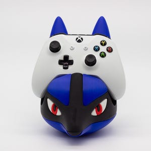 Puede incluir: Un mando de Xbox blanco y azul descansa sobre una base azul, negra y roja con temática de personaje. El mando tiene botones negros y una cruceta. La base tiene ojos rojos y orejas azules puntiagudas.