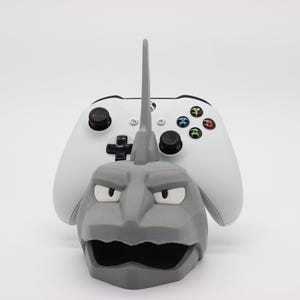 Puede incluir: Un soporte gris impreso en 3D con el tema de Onix con un mando Xbox blanco. El soporte presenta la cara del personaje Pokémon y una estructura alta y puntiaguda. El mando tiene botones negros y botones de acción coloridos.