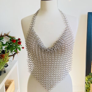 May include: A silver chainmail halter top displayed on a mannequin. The top is made of small, interlocking metal rings, creating a shimmering, textured surface. The halter design features a triangular shape that covers the chest.