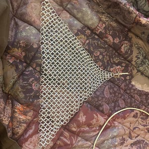 May include: Silver chainmail top in a triangular design. The chainmail is constructed from small, interconnected metal rings. The top is presented on a floral patterned fabric.