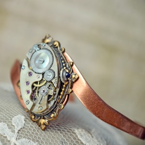 May include: A copper cuff bracelet with a vintage watch face attached. The watch face has a silver finish and features intricate gears and pink jewels.