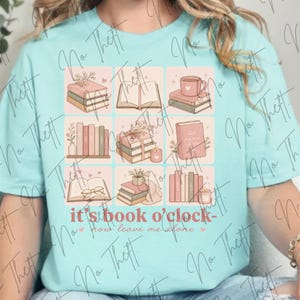 Może przedstawiać: Jasnoturkusowy t-shirt z siatką dziewięciu ilustracji przedstawiających książki, kubki i świece w delikatnych pastelowych odcieniach. Pod siatką znajduje się napis "it's book o'clock- now leave me alone" czerwonawą czcionką z małymi serduszkami.