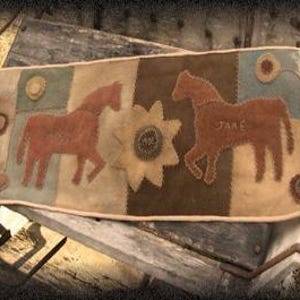 May include: A rectangular, patchwork-style textile featuring two stylized horses in reddish-brown, flanking a large, tan sunflower. The textile includes patches of brown, blue, and tan, with the name "Jake" stitched on one horse. The textile is resting on weathered wooden planks.