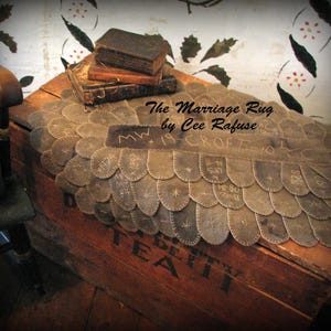 Puede incluir: Un cofre de madera marrón vintage con las palabras "BETTY TEA" estampadas en el lateral. Una alfombra decorativa marrón envejecida con bordes festoneados y detalles cosidos está drapeada sobre el cofre. La alfombra tiene el texto "The Marriage Rug by Cee Rafuse".