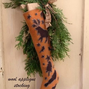 May include: An orange wool applique Christmas stocking with dark blue stitched designs of a moose, trees, and stars. The stocking is decorated with greenery and the text "The Medway Stocking" and "wool applique stocking designed by Cee Rafuse".