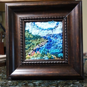 May include: A framed textile art piece depicting a landscape scene. The artwork features a textured, colorful design with shades of blue, green, and white, framed in a dark brown, ornate wooden frame. The frame has a beaded inner edge.