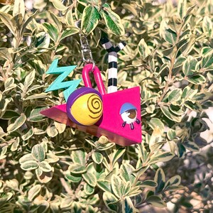 May include: A whimsical keychain featuring a pink geometric design with a yellow and purple eye-like shape, a blue zig-zag, and a small astronaut figure. The keychain also includes a black and white striped element and a silver-colored chain.