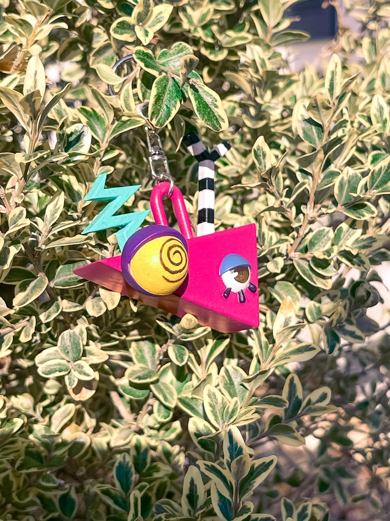 May include: A whimsical keychain featuring a pink geometric design with a yellow and purple eye-like shape, a blue zig-zag, and a small astronaut figure. The keychain also includes a black and white striped element and a silver-colored chain.
