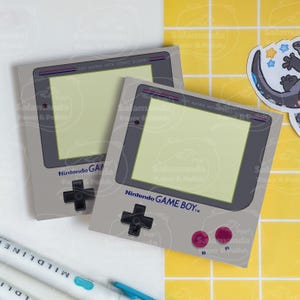 May include: Two gray notepads designed as Nintendo Game Boy consoles. Each notepad features a light green screen, a black directional pad, and pink buttons. The text "Nintendo GAME BOY" is printed on the front. The background is white and yellow.