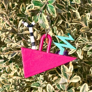 May include: A vibrant pink triangle-shaped keychain with a silver chain. It features a black and white striped palm tree, a turquoise zig-zag design, and a pink handle. The keychain is set against a backdrop of green and yellow foliage.