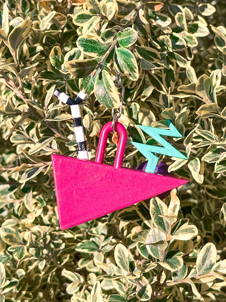 May include: A vibrant pink triangle-shaped keychain with a silver chain. It features a black and white striped palm tree, a turquoise zig-zag design, and a pink handle. The keychain is set against a backdrop of green and yellow foliage.