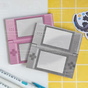 Può includere: Due blocchi per appunti decorativi a forma di Nintendo DS. Uno è rosa, l'altro grigio. Entrambi hanno uno schermo bianco e il classico layout dei pulsanti e del D-pad. Un adesivo di una salamandra è sullo sfondo.