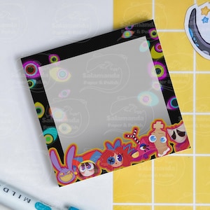 May include: A square notepad with a black border featuring colorful cartoon faces and eye designs. The notepad has a transparent overlay with the text "Salamanda Paper & Polish". A sticker with a star and octopus is visible.