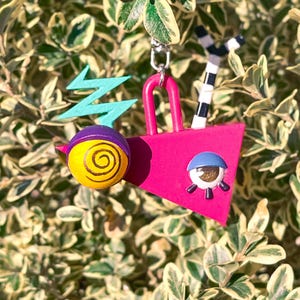 May include: A whimsical, colorful charm featuring a pink geometric shape with a blue eye, a yellow and purple ball with a spiral design, and a teal lightning bolt. A black and white striped element and a silver chain complete the design.