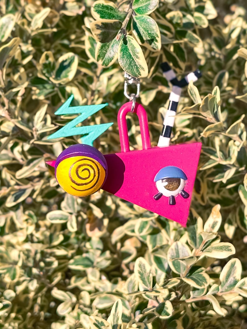 May include: A whimsical, colorful charm featuring a pink geometric shape with a blue eye, a yellow and purple ball with a spiral design, and a teal lightning bolt. A black and white striped element and a silver chain complete the design.