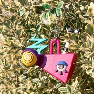 May include: A whimsical keychain featuring a pink padlock shape with an eye design, a yellow and purple snail, and a teal lightning bolt. The keychain is attached to a silver chain and is set against a backdrop of green and yellow foliage.