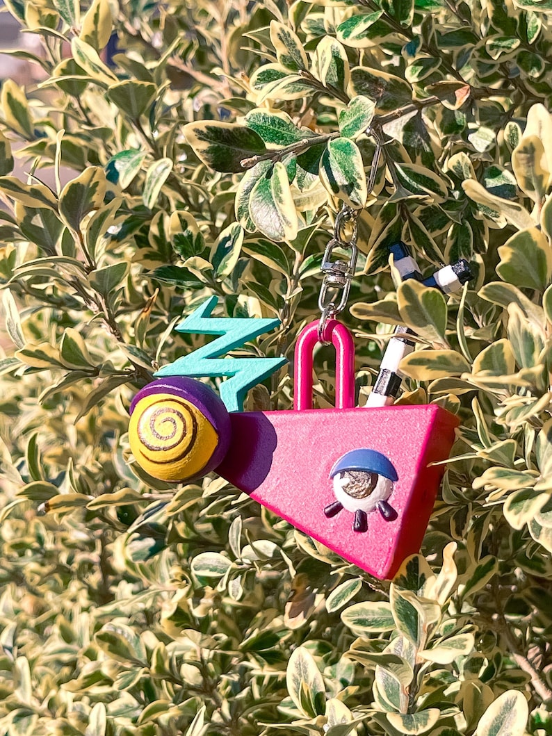 May include: A whimsical keychain featuring a pink padlock shape with an eye design, a yellow and purple snail, and a teal lightning bolt. The keychain is attached to a silver chain and is set against a backdrop of green and yellow foliage.