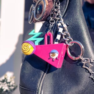 May include: Black leather handbag with decorative charms. Charms include a pink triangle, a teal zigzag, and a yellow and purple orb. The bag has silver chain accents and a brown suede detail.