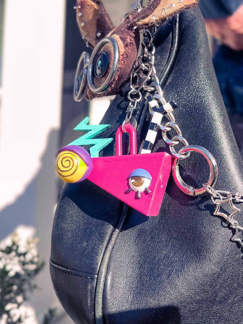 May include: Black leather handbag with decorative charms. Charms include a pink triangle, a teal zigzag, and a yellow and purple orb. The bag has silver chain accents and a brown suede detail.