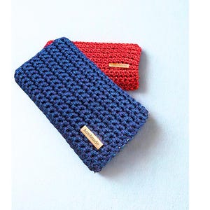 Könnte beinhalten: Zwei handgefertigte Häkeltaschen. Eine ist tiefblau mit einem Holzschild „handmade“, die andere rot. Die Taschen sind rechteckig und haben ein strukturiertes, gewebtes Aussehen. Der Hintergrund ist hellblau.