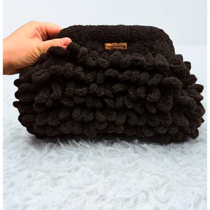 May include: A dark brown, handmade clutch bag with a textured, looped design. The bag has a rectangular shape and a small, rectangular tag with text. The bag is held by a hand, and the background is a white, fluffy surface.