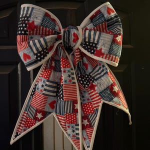 Pode incluir: Um grande laço decorativo com um padrão de patchwork em vermelho, branco e azul. O tecido apresenta vários desenhos, incluindo estrelas, riscas e corações, com debrum branco ao longo das bordas do laço e das suas duas longas pontas.