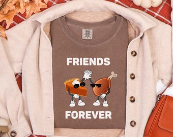 Può includere: Una maglietta marrone con le parole "FRIENDS FOREVER" in bianco. La maglietta presenta un'immagine a cartoni animati di una fetta di torta e una coscia di tacchino arrosto con occhiali da sole e che si battono il cinque. La maglietta è sovrapposta a una giacca beige.