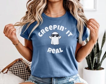 May include: A blue t-shirt with the text "CREEPIN' IT REAL" in white, featuring a cartoon ghost wearing sunglasses. The shirt is short-sleeved and has a round neckline. The overall theme is Halloween.