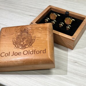 May include: A wooden box with an engraved crest and the text "Col Joe Oldford". The box is open, displaying gold cufflinks and shirt studs on a black velvet lining. The box is made of light-coloured wood.