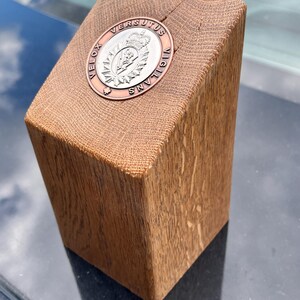 Könnte beinhalten: Ein Holzblock mit einem Metallabzeichen. Das Abzeichen hat ein kreisförmiges Design mit den Worten "VELOX VERSUTUS VIGILANS" um ein zentrales Wappen. Das Holz hat eine natürliche Maserung und eine warme braune Farbe.