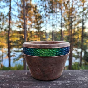 Può includere: Una ciotola di legno con una fascia di rame e un intarsio di vetro strutturato blu e verde. La ciotola è posta su uno sfondo sfocato di alberi e un lago.