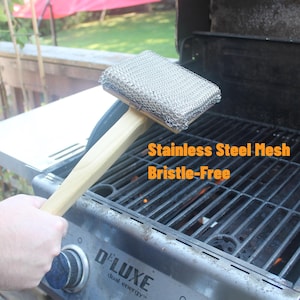 Puede incluir: Un cepillo para parrilla de malla de acero inoxidable con mango de madera. El cepillo no tiene cerdas y está diseñado para limpiar las rejillas de la parrilla. El texto "Stainless Steel Mesh Bristle-Free" es visible.