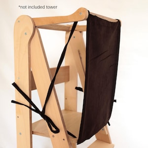 May include: A wooden learning tower with a brown safety net attached. The text "LEAVING TOWER SAFETY NET" is printed at the top. Black ties secure the net to the tower frame. The tower is made of light-coloured wood.