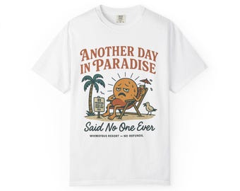 May include: White t-shirt with a humorous graphic. The design features a sun-like character in a beach chair, with the text "ANOTHER DAY IN PARADISE Said No One Ever." A palm tree and a seagull are also included in the illustration.