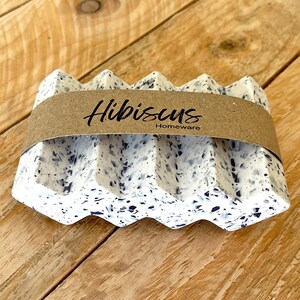 Puede incluir: Un jabonera moteada blanca y negra con un diseño acanalado. La jabonera tiene una banda de papel marrón con la palabra "Hibiscus" y "Homeware" impresa. La jabonera está sobre una superficie de madera.