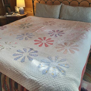 May include: A white quilt with a floral pattern in shades of pink, blue, and red. The quilt is on a bed with a wooden headboard and a white and blue pillow.